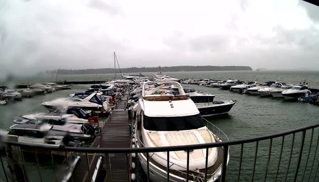 A view of a marina under overcast skies, with several boats docked in the water. The image appears slightly blurred, likely due to rain. In the foreground, a large white boat is visible, with a silver railing in front. Surrounding it are various other boats of different sizes and designs, some with canopies. Beyond the boats, a shoreline with trees can be seen in the distance. The overall atmosphere is calm, but the weather looks gloomy.