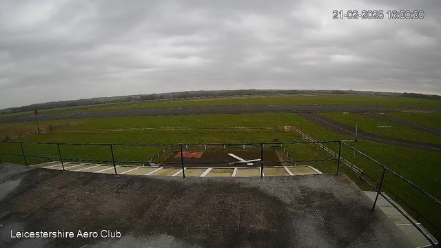 A cloudy sky fills the top portion of the image. Below, there is a green grassy field that stretches across the center, with a runway made of asphalt running horizontally. On the left, a small red and white traffic light tower stands next to the edge of the field. There is a wooden fence marking the boundaries of the area. The lower part of the image shows a flat rooftop with visible panels, and there are a few scattered objects on the ground, including a white piece of material. The image is timestamped 21-02-2025 at 16:00:20 and labeled with "Leicestershire Aero Club" at the bottom left.