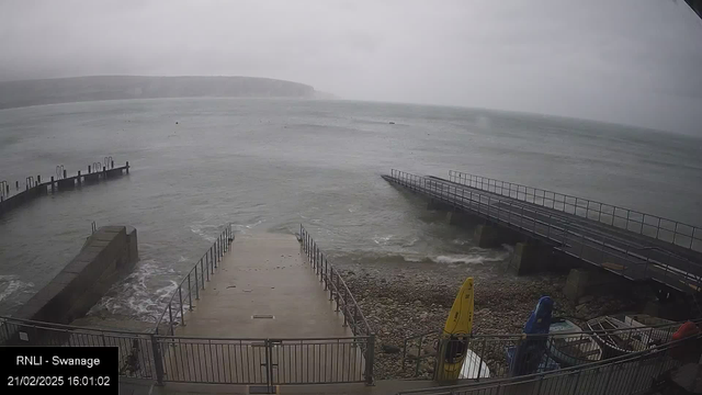 A cloudy and overcast scene at a waterfront location. In the foreground, a concrete pathway leads down to the water, lined with a metal railing. To the left, a small stone structure juts out towards the sea. In the background, boats are visible on the water, which appears choppy. To the right, a longer wooden pier extends out into the water. On the shore, several boats in various colors are positioned along a rocky area. The sky is grey and the atmosphere feels tranquil yet moody.