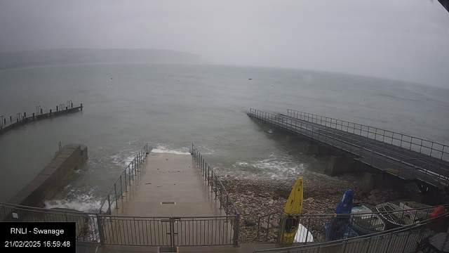 A coastal scene showing a cloudy sky over the sea. The water is calm with small waves reaching the shore. A set of stone steps leads down to the water, flanked by metal railings. To the right, there is a wooden pier extending into the sea. Various boats are tied up along the pier and one yellow kayak is visible on the left side near the rocks. The shoreline is rocky, and the overall atmosphere is overcast and serene.