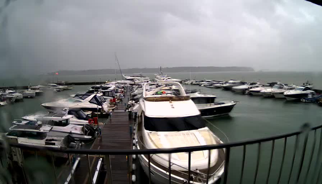 A view of a marina filled with various boats, some on a floating dock and others moored in the water. The scene is overcast with dark clouds, indicating a stormy weather condition. Water appears choppy, and there are several boats of different sizes, mostly in white and gray colors. The image captures a sense of calm amid potential rough weather, with the distant shoreline visible in the background.