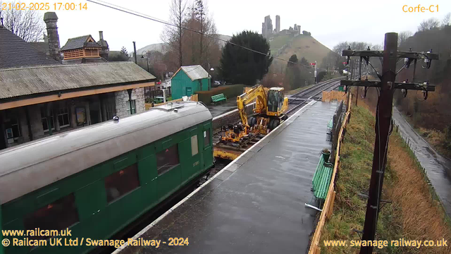 A train with a green exterior is parked on a wet platform at a railway station. To the right, a yellow construction vehicle is positioned on the tracks, possibly for maintenance work. In the background, there are trees, benches, and a small green building. A hill can be seen in the distance, with ruins of a castle atop it. The sky is overcast, indicating a gloomy weather condition.