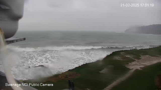 The image shows a coastal scene with gray skies and rough waves crashing against the shore. In the foreground, there is a grassy area, and a pathway leads towards the shore. The water is choppy, indicating stormy weather. The scene appears to be taken from a high vantage point, possibly from a webcam. A timestamp at the top indicates the date and time of the capture.