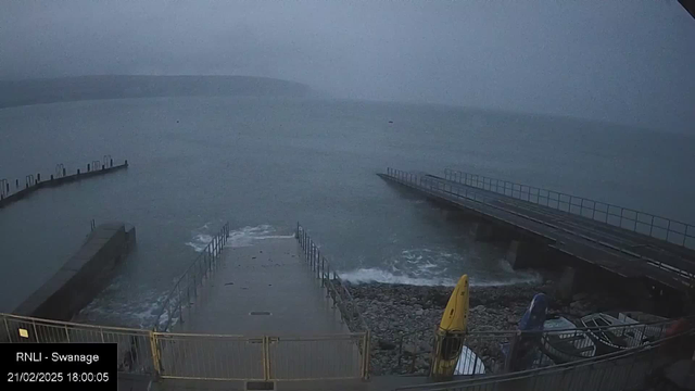 A grey and overcast scene of a waterfront area, featuring a concrete pier leading into the water. Waves can be seen lapping against the shore, with a rocky shoreline on the right side. There are two boat ramps, one with a yellow kayak aligned on it. An additional, larger wooden pier extends into the water on the left. In the background, a misty landscape of hills can be seen fading into the distance. The overall atmosphere appears calm yet slightly gloomy, suggesting an overcast day.