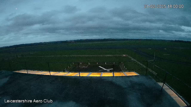 A view from a webcam at Leicestershire Aero Club showing a cloudy dusk sky and a grassy airfield. In the foreground, there is a platform with yellow markings and a white cross symbol on the ground. The background features a wide expanse of fields, with a railing visible along the edge of the platform. The atmosphere is muted with gray clouds overhead, indicating the evening time.