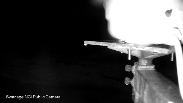 A dimly lit image showing part of a mounting bracket for a camera, with metal components and a blurred, glowing area in the background. The overall scene appears shadowy, indicating low visibility, with the watermark for "Swanage NCI Public Camera" in the lower left corner.