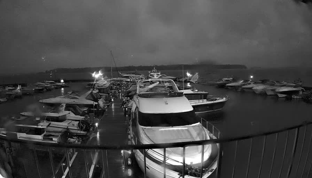 A dark, cloudy scene of a marina filled with various boats. The image is in black and white, with several yachts and smaller boats docked near a wooden platform. Soft lighting from the boats creates reflections on the water's surface. The atmosphere appears calm but dim, suggesting early morning or late evening conditions.