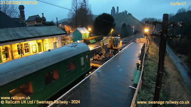 A train station scene at dusk with a green train car on the left side. In the foreground, a yellow construction vehicle is on the railway tracks, working on maintenance. A stone building with illuminated windows is in the background, along with a green shed and benches. In the distance, a hill is visible with ruins on top. The atmosphere appears calm and slightly overcast, with streetlights providing soft illumination.