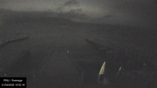 A dimly lit seaside view showing a horizon line where the sea meets the sky. There is a faint outline of a pier extending into the water on the left, with a few boats visible at the edge. The scene is shrouded in darkness, with cloudy skies above, giving a moody atmosphere. The date and time in the bottom left corner indicates it was taken at night.