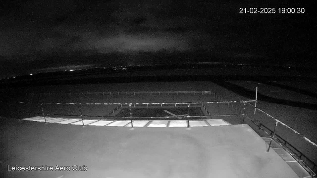 A dark image captured by a webcam showing the Leicestershire Aero Club. The foreground features a flat surface with a railing and several panels. In the background, faint lights are visible, indicating other structures or vehicles at a distance. The sky is cloudy, and the overall scene is predominantly dark, with limited visibility. The timestamp in the corner reads 21-02-2025 19:00:30.