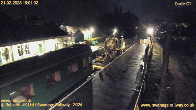 A nighttime image of a train station under dim lighting. In the foreground, a green train is parked on the platform. To the right, there is a construction machine working on the tracks, with lights illuminating the area. A row of benches can be seen along the platform. In the background, the station building has large windows and appears to have a green door. The scene is primarily dark, with various light sources highlighting certain areas.