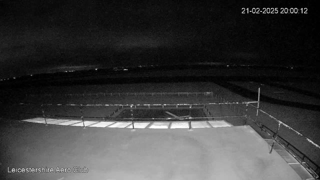 A dark night scene captured from a webcam at the Leicestershire Aero Club. The foreground shows a flat rooftop with a railing and a rectangular opening. In the background, faint city lights are visible against a dark sky. The timestamp in the top right corner reads "21-02-2025 20:00:12."