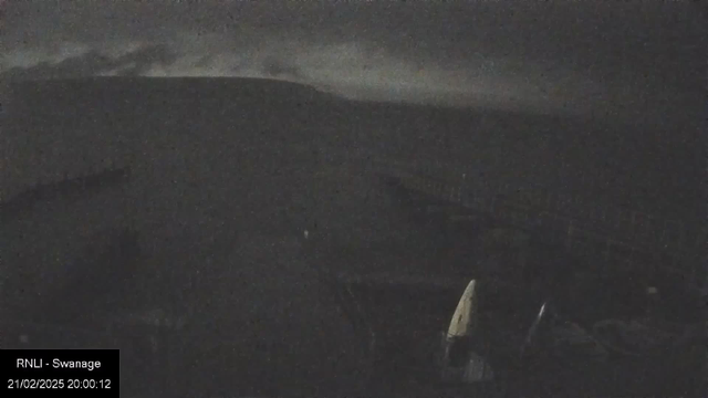 A dark, cloudy sky fills the top part of the image, with hints of lighter clouds in the distance. Below, a silhouette of land is visible, presumably a coastline or hillside. The foreground features indistinct shapes, possibly boats or structures, blending into the darkness. The overall scene has a dim and moody atmosphere, typical of nighttime or low-light conditions.