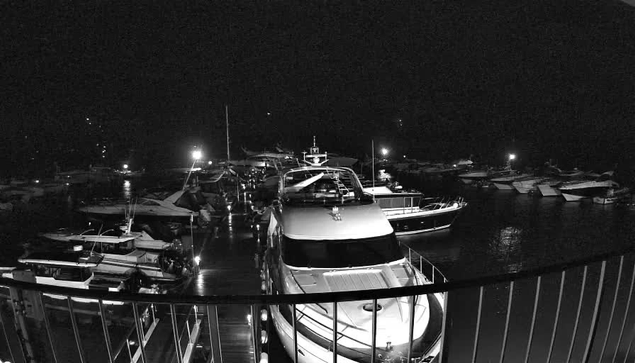 A black and white image of a marina at night, featuring multiple boats and yachts docked along the water. The scene is illuminated by various lights reflecting off the water's surface. In the foreground, a large yacht is visible, with its structure prominently highlighted. The surrounding boats appear smaller and are arranged in the background, creating a sense of depth in the composition. Some lights from the marina are scattered throughout the scene, adding to the night ambiance.