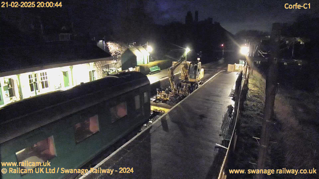 A dimly lit railway station scene at night showing a green train car on the left side of the image. In the center, a yellow construction vehicle is positioned on the platform, working on the track. To the right, there are benches along the platform, illuminated by a nearby light. The background features a few buildings and trees, slightly visible in the darkness. The overall atmosphere is calm and work-related.