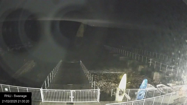 A dark scene captured from a webcam, showing a pier with a wooden structure and a rocky shoreline. The area is illuminated by a faint light. There are two surfboards, one yellow and one blue, positioned upright against a nearby railing. The image has a slight blur and some water droplets, indicating moisture on the camera lens. No people are visible in the scene.