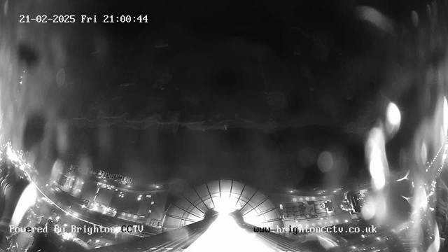 Black and white image from a webcam showing a dimly lit area with faint outlines of buildings or structures at the bottom. The scene is mostly dark with some bright, reflective surfaces, possibly lights, visible towards the bottom. The timestamp in the top left corner indicates the date and time as 21-02-2025, Friday, 21:00:44. The text at the bottom left reads "Powered By Brighton CCTV," while the bottom right displays the website URL "www.brightoncctv.co.uk."