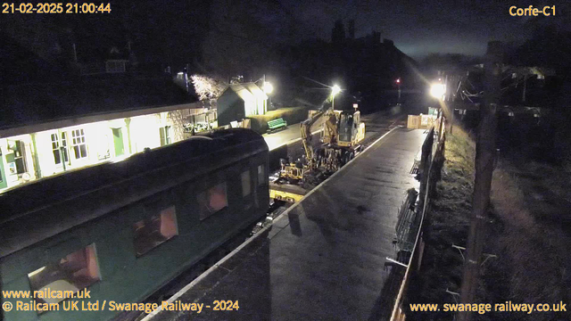 A dimly lit railway station at night. On the left, a green train carriage is partially visible. In the center, a construction vehicle is working on the tracks. The platform is empty, with a few benches along the side. On the right, a signal light is illuminated. Some trees and a low building are in the background, and the atmosphere is quiet and still.