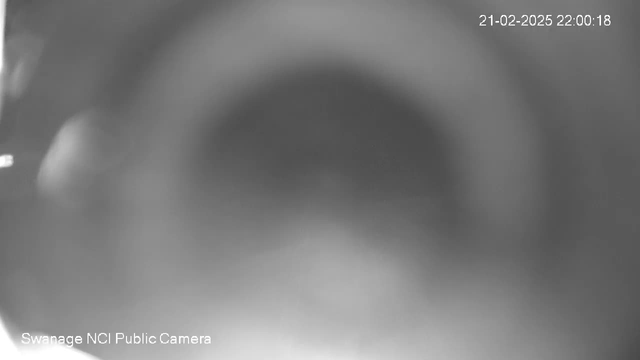 A blurry, monochrome image showing a circular gradient that appears darker in the center and lighter towards the edges. There are faint outlines of shapes possibly indicating objects near the left edge. The timestamp in the upper right corner reads "21-02-2025 22:00:18," and the bottom left indicates "Swanage NCI Public Camera."