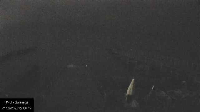 A dark webcam image showing a blurred and indistinct scene, possibly of a coastal area at night. The outlines of a pier or boardwalk can be faintly seen in the background, along with some indistinguishable shapes in the foreground. The overall lighting is low, contributing to the lack of detail in the image. In the lower left corner, there is a timestamp and location label indicating it is from Swanage.