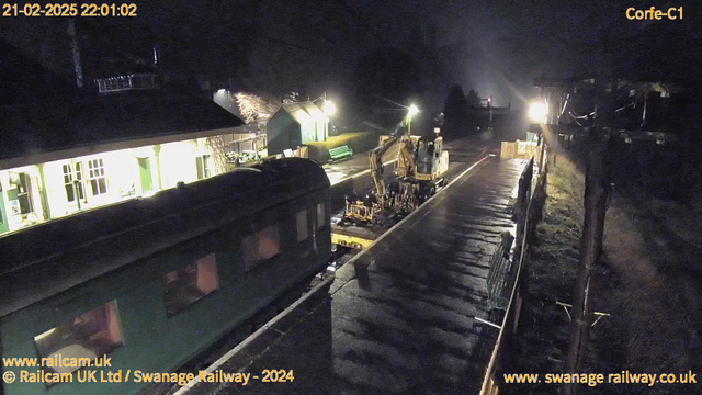 A dimly lit railway station at night shows a green train car on the left side and a yellow construction vehicle working on the tracks. The station building has multiple windows, some illuminated, and features a green wooden bench nearby. In the background, there are additional structures and faint lights. The wet platform reflects the light, and the atmosphere appears quiet and somewhat secluded.