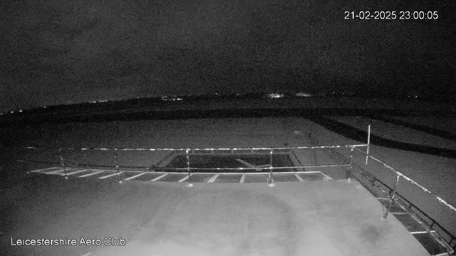 A dark night scene captured from a webcam at Leicestershire Aero Club. In the foreground, there is a railing along a flat surface, likely a rooftop or balcony, with several vertical posts. Below the railing, a square area is visible, possibly an opening or enclosed space. The background features a flat area that appears to be an airfield under dim lighting, with distant lights twinkling in the distance, suggesting other structures or the horizon. The time displayed in the corner shows 23:00:05 on February 21, 2025.