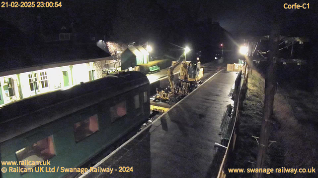 A nighttime scene at a railway station. The image shows a train carriage in dark teal parked on the left side. In the foreground, there is construction machinery, including a yellow digger, working on the platform. The platform is illuminated with lights, and a building with large windows and green doors is visible in the background. There are benches along the platform and some trees. The overall atmosphere is quiet and dimly lit, typical of a late evening setting.