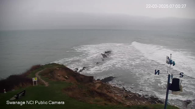 A coastal scene captured from a webcam shows a cloudy sky over a calm sea. Gentle waves lap against rocky formations jutting out into the water. In the foreground, there is a patch of grassy land with a few benches and a sign. A weather vane with cardinal directions is visible on the right. The image appears slightly overcast, indicating a cool atmosphere.