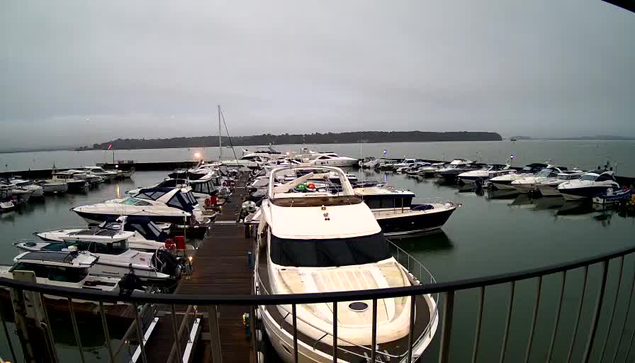 A marina scene with numerous boats docked in calm water. The sky is overcast, giving a grayish tone to the image. Several motorboats are visible, some with white and blue colors, along with various accessories and equipment on board. A wooden walkway leads along the edge of the marina, and there are mooring poles visible. The overall atmosphere appears quiet and serene.