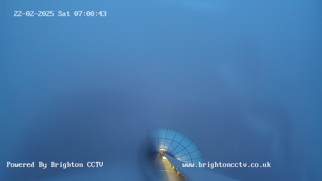 A foggy scene from a webcam shows a circular, glass-covered structure leading into the distance. The environment is predominantly blue with a few soft shapes appearing vague in the background. A faint glow from lights illuminates the pathway inside the structure, suggesting early morning hours. The date and time are displayed in the upper left corner.