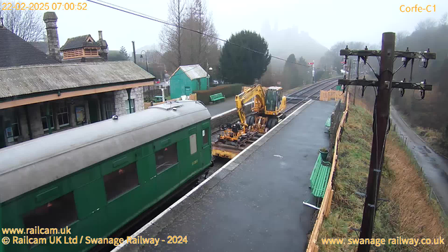 A railway station scene shrouded in mist. In the foreground, a green train car is parked along the platform. To the right, a yellow excavation machine is positioned on the tracks. There are wooden benches painted green along the platform. In the background, a stone building with a peaked roof is visible, along with a green shed-like structure. A telephone pole with wires stands on the edge of the platform, and a winding road is seen curving away into the fog. The setting appears quiet and somewhat deserted.