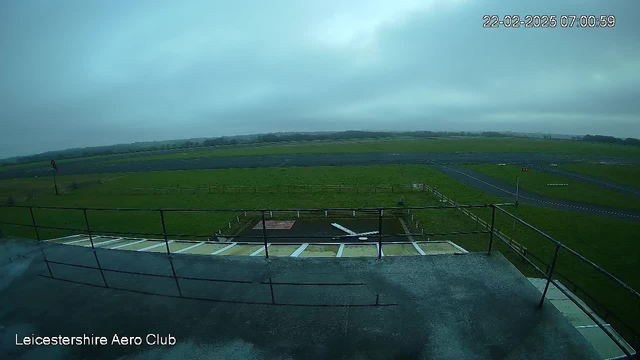 A view from a webcam atop a building at Leicestershire Aero Club. The image shows a cloudy sky with limited visibility. In the foreground, there is a rooftop railing and a paved area. Below, an expansive green field stretches out, intersected by a dark runway and a few white markings. In the distance, low hills are visible through the haze. The scene has a tranquil atmosphere, indicating early morning.