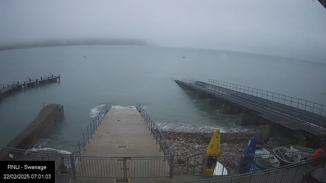 A view of a foggy waterfront with a mist-covered sea. In the foreground, there is a concrete jetty leading to the water, flanked by metal railings. To the right, several colorful kayaks are resting on stony ground. In the background, there are wooden piers extending into the water. The overall atmosphere is calm but lacks visibility due to fog.