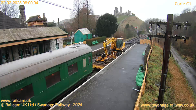 A railway station scene with a green train car parked on the left side. Next to it, there is yellow construction machinery on a flatbed, positioned toward the tracks. In the background, there are grassy slopes leading up to ruins of a castle on a hill. The station platform is mostly empty, with green wooden benches along the side. The setting is overcast and a bit damp, typical of early morning. Utility poles with wires run along the platform.