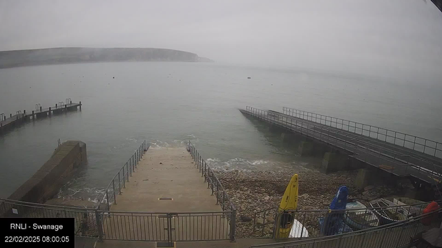 A foggy seaside scene shows calm waters with a distant shoreline and hills. There are two piers extending into the water, one on the left with a series of posts and the other on the right with a flat surface. A wide staircase leads down to the water, surrounded by rocky ground. Several kayaks in bright colors, including yellow and blue, are secured near the water's edge. The sky appears overcast, creating a muted atmosphere.