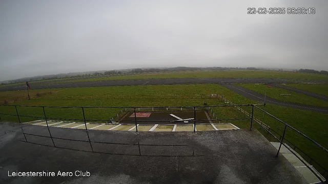 A view from a webcam overlooking a wide, green field with a tarmac runway. The sky is cloudy and gray, indicating overcast weather. In the foreground, there is a railing along a flat surface, with a small area on the ground marked by a red square. To the left, a flagpole stands with a flag. The landscape extends to the horizon, featuring a few low hills and a fence bordering the field. The scene appears quiet and still, typical of an early morning at an airfield.