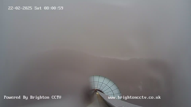 A foggy landscape is seen from a webcam perspective. The scene is blurred and mostly featureless, with a curved roof structure at the bottom suggesting an observation point or architectural element. The date and time read "22-02-2025 Sat 08:00:59" at the top of the image, and a watermark indicates "Powered By Brighton CCTV" at the bottom left, and a web address on the bottom right.