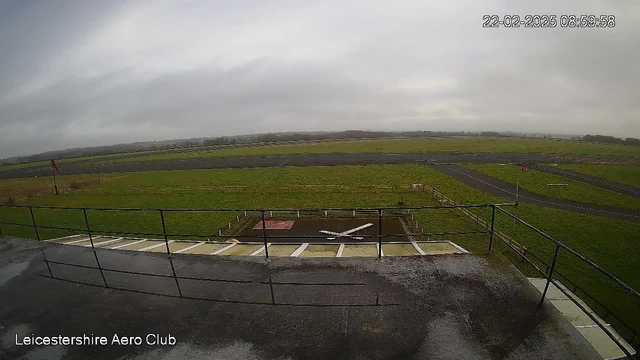 A cloudy sky is visible above a grassy airfield. In the foreground, there is a railing that suggests a viewing area. Below the railing, a small white airplane is parked on a runway that has a red and white marked area. The runway expands into the distance, bordered by green grass and fences. A windsock is visible on the left side of the image, indicating wind direction. The scene appears calm with no visible aircraft in flight.