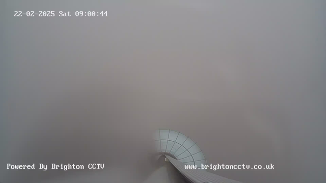 A foggy view from a webcam showing a curved white structure that leads out of the frame. The background is mostly gray, indicating low visibility. The image displays the date and time at the top and a watermark at the bottom indicating it is powered by Brighton CCTV.