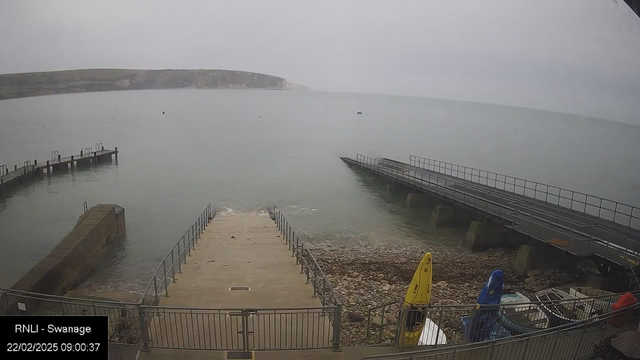 A calm seaside view under a cloudy sky. In the foreground, a concrete ramp leads down to the water, flanked by a rocky shoreline. To the left, a wooden pier extends into the water, and on the right, a metal ramp ends at the water's edge. Several boats, including yellow and blue kayaks, are stored along the shore. The sea appears still, with a few floating objects visible. The date and time are displayed in the corner of the image.