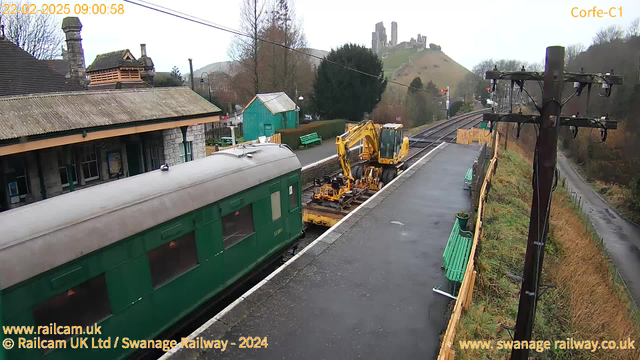 A view of a railway station with an old green train parked on one side. In the foreground, there is a yellow construction machine positioned next to the train, working on the tracks. The platform is wet, likely from recent rain. Behind the station, there are trees, and in the distance, a historic castle ruin can be seen atop a hill. There are green benches on the platform, and a few small buildings, including one in teal color. The image is taken during daylight, with overcast sky conditions.