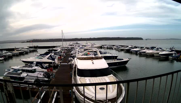 A marina scene featuring numerous boats docked in the water. In the foreground, a white boat with a circular window and a green canopy is visible. Other boats of various sizes and colors surround it, some with blue and white stripes. The water is calm, reflecting the cloudy sky above, with scattered clouds and a hint of light. In the background, there are trees and land in the distance. A wooden dock is partially visible along the bottom of the image, leading to the water.