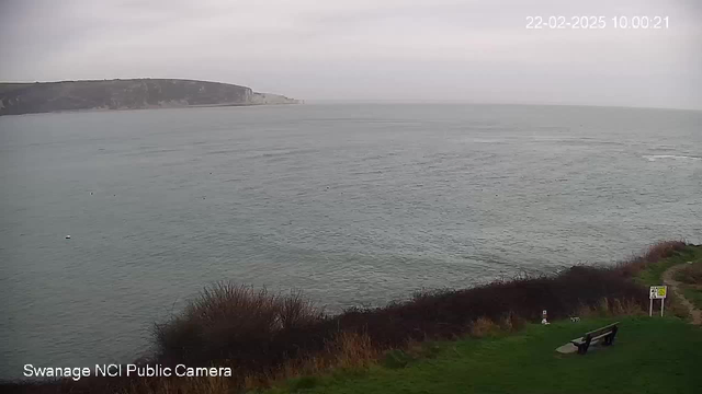 A cloudy view of a calm sea with gentle waves. The coastline is visible in the background, showcasing rugged cliffs. In the foreground, there is a grassy area with some bushes. A wooden bench is positioned to the right, and a sign can be seen nearby, likely providing information or warnings. The scene appears tranquil and scenic.