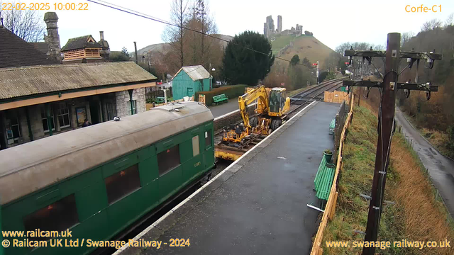 A railway station scene with a green train carriage on the left, partially visible. Next to it, construction equipment is parked on the platform. In the background, there are stone structures on a hill, likely ruins, and trees are visible. The weather appears cloudy, and there are benches along the platform. A road is seen on the right side, with some grass and trees bordering it. The image is timestamped and labeled with the location "Corfe-C1."