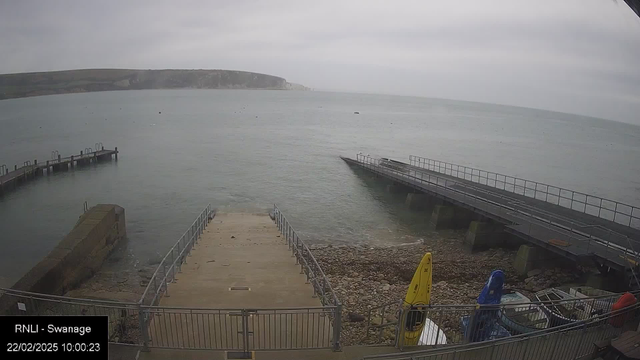 A view of a rocky shoreline with a calm sea in the foreground. There are two docks extending into the water: one is a wooden pier on the left, and the other is a wider, metal structure on the right. Various boats are visible parked along the docks, including a yellow kayak and a blue one. The sky is overcast, with a light gray color, and a few distant cliffs can be seen in the background along the horizon. The time and date are displayed in the lower left corner.