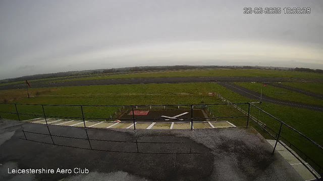 A view from a high point overlooking an airfield. The foreground features a railing and part of a flat surface, which appears wet. Below is a grassy field that stretches across the image, bordered by a runway in the background that leads off into the distance. There are a few red flags on poles, and a white painted "X" marked on the ground. The sky is overcast, suggesting a cloudy day. A timestamp shows the date and time: February 22, 2025, at 10:00:38.