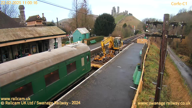 A view of Corfe Castle railway station, featuring a green train carriage on the left side of the image. Next to the train, construction equipment, including a yellow excavator, is parked on the platform. In the background, there is a hillside with the ruins of Corfe Castle atop it, surrounded by trees and a partly cloudy sky. The station building, showing a stone facade and a sloped roof, can be seen on the left. Wooden benches and a green shed are situated in the vicinity. The image captures a quiet moment at the station with no passengers visible.