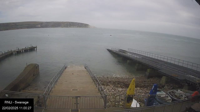 A view of a coastal area featuring a calm sea under a cloudy sky. In the foreground, there are steps leading down to the water, bordered by a metal railing. To the left, a small jetty extends into the water. On the right, a larger structure juts out, also surrounded by a railing. There are several small boats near the shore, including two yellow and blue kayaks positioned on the ground. In the distance, the coastline is visible with a rocky cliff. The scene appears tranquil and overcast. The date and time in the corner indicate it is 11:00 AM on February 22, 2025.