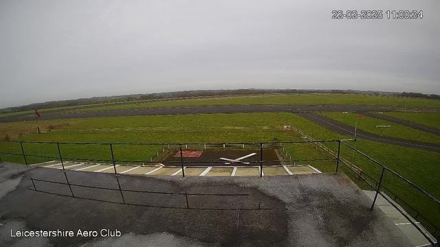 A view from a webcam at Leicestershire Aero Club shows a grassy airfield under a cloudy sky. In the foreground, there is a section of a building's balcony railing, and below it, a small area with a white aircraft silhouette on the ground. The airfield is bordered by a fence, and further in the distance, there are runways and scattered flags. The scene is primarily green with shades of gray from the sky.