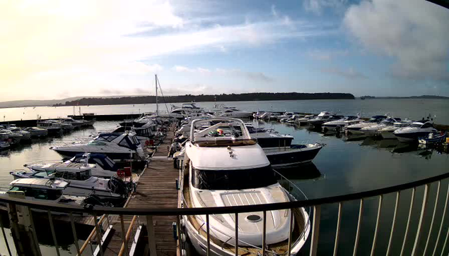 A marina scene with numerous boats docked in calm water. Several motorboats and yachts are aligned at the pier, while a wooden walkway runs alongside them. The background features a cloudy sky and a distant shoreline with trees. The overall atmosphere is tranquil and nautical, evoking a sense of leisure by the water.